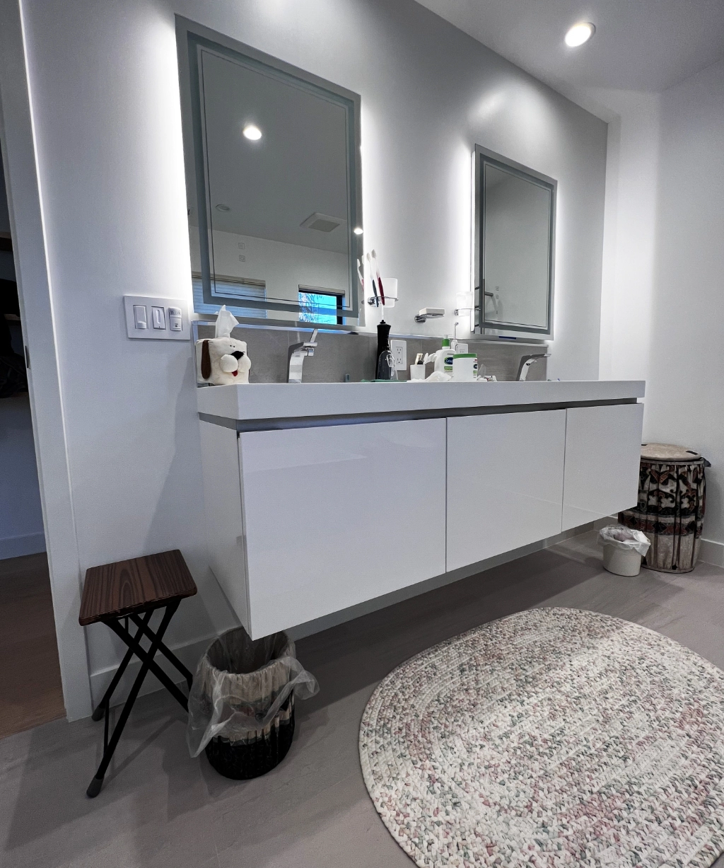 A modern bathroom with a sleek double sink vanity, two mirrors, a small stool, and a round rug on the floor. Toiletries and a decorative item are placed on the counter. Ideal for those seeking luxury home remodeling services to elevate their space.
