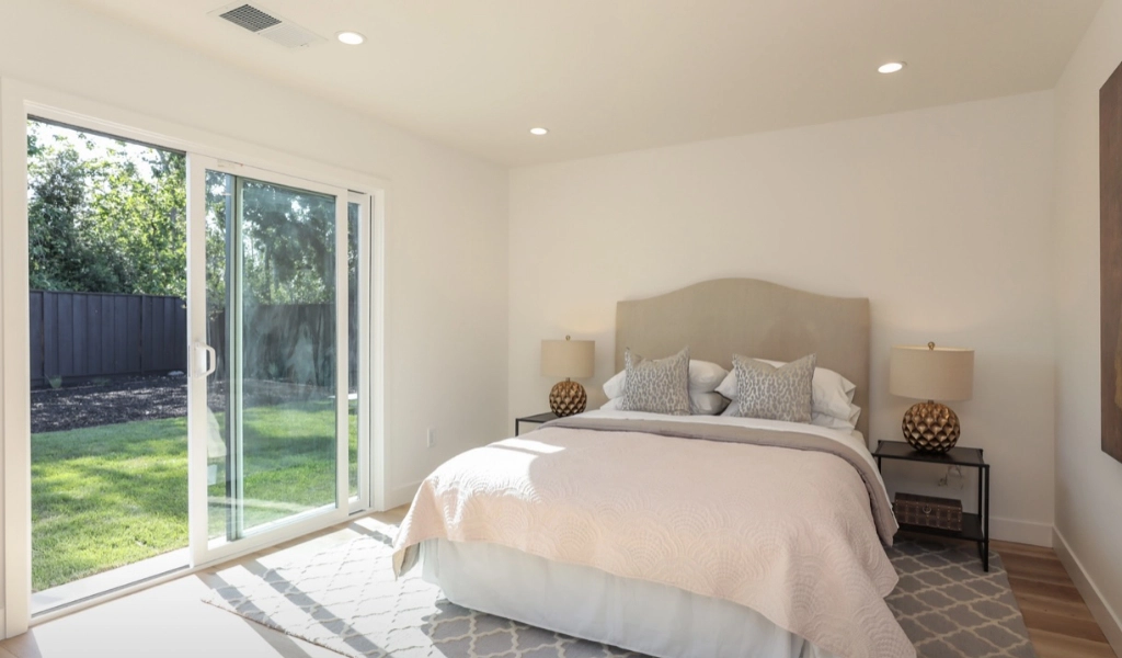 A bedroom crafted by a home remodeling company features a large bed with two lamps on side tables, a sliding glass door leading outside, and a white rug on wooden flooring. The walls are white, adorned with a framed picture to the right, embodying elegance and style.