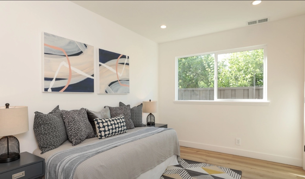 A modern bedroom featuring a bed, abstract artwork above, and two lamps, with a window revealing lush greenery outside. The light walls and wood flooring create an inviting space, perfect for those considering home remodeling services to enhance their living environment.