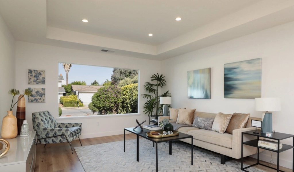 A modern living room with a beige sofa, patterned armchair, and two abstract paintings is the epitome of elegance after consulting a top home remodeling company. A large window showcases greenery, while a geometric rug adds flair beside decorative items on the coffee table.