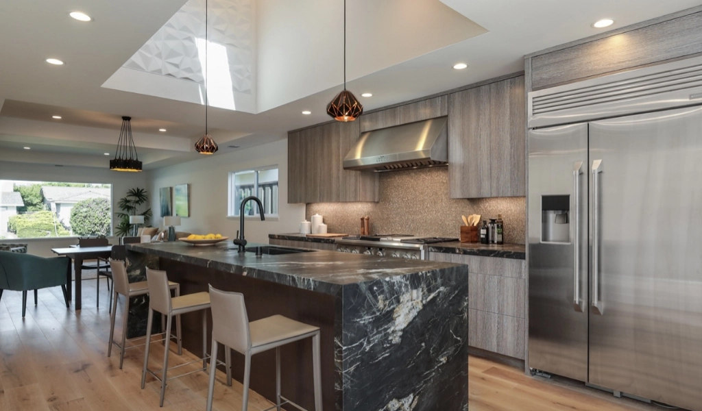 Modern kitchen with a marble island, bar stools, stainless steel appliances, and pendant lighting—a testament to expert home remodeling by a skilled contractor. The dining area is elegantly visible in the background.