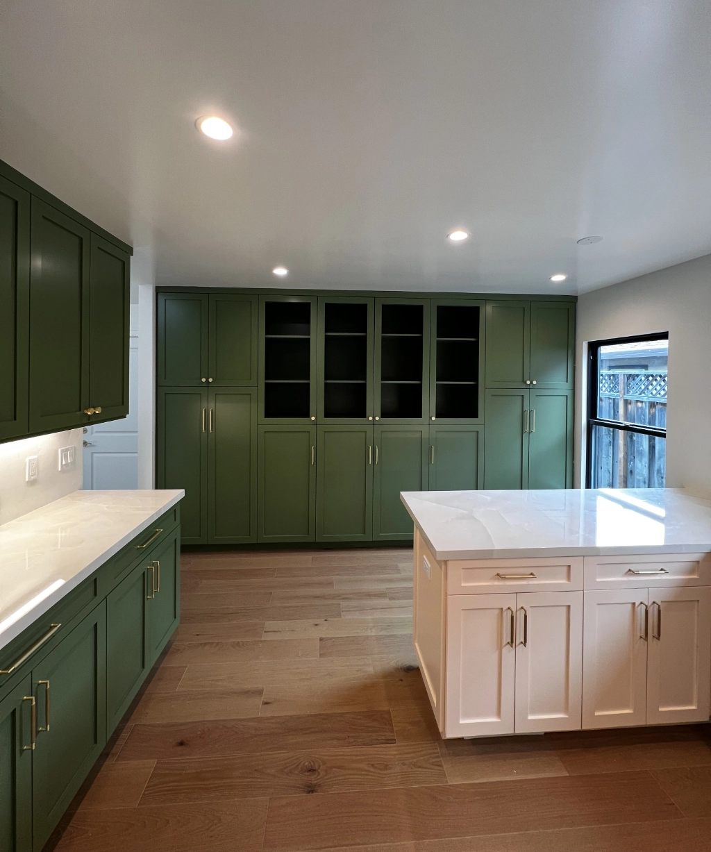 A modern kitchen makeover features green cabinets, a large white island, and wood flooring. Natural light pours in through a window on the right. This stylish space is crafted by an expert home remodeling contractor.