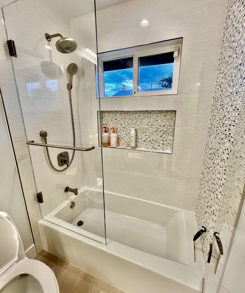 This modern bathroom, perfect for any home remodeling project, features a white bathtub, glass shower doors, and a sleek showerhead. A built-in niche holds toiletries neatly, while a small window above the tub provides a charming view outside. Consider partnering with a trusted home remodeling contractor to create yours.