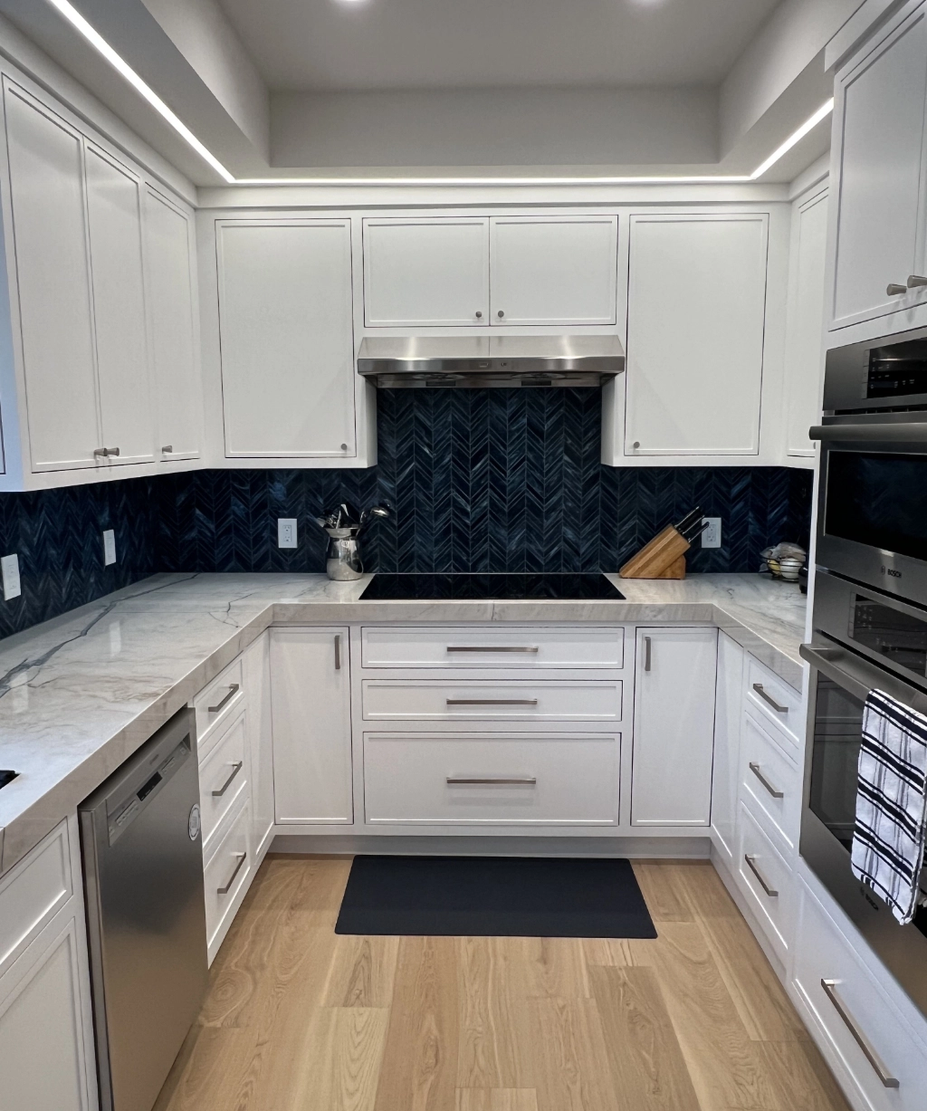 Modern kitchen with white cabinets, marble countertops, and a dark blue herringbone tile backsplash. Stainless steel appliances gleam under the sleek design, while wooden flooring adds warmth. Perfectly crafted for those who appreciate luxury home remodeling services.