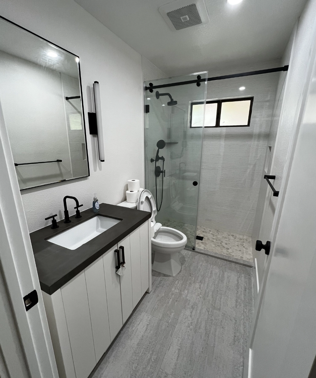 Modern bathroom designed with a sleek touch, featuring a glass shower, black fixtures, and a white vanity with a black countertop. A wall-mounted mirror and toilet complement the space. Light gray floor tiles and a small window complete this home remodeling contractor's masterpiece.