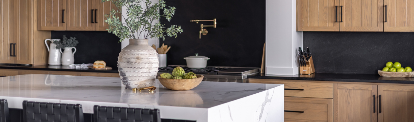 Modern kitchen with wooden cabinets, a marble island featuring a decorative vase and fruit bowls, and a stove with a brass faucet against a black backsplash. Perfect for showcasing your home remodeling services.