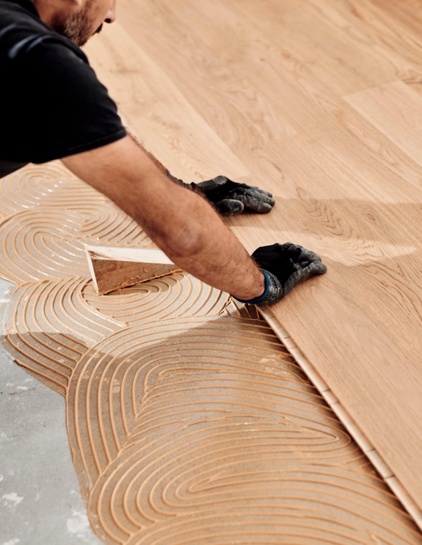 Person installing wooden flooring with adhesive on a concrete surface, wearing black gloves and a black shirt.