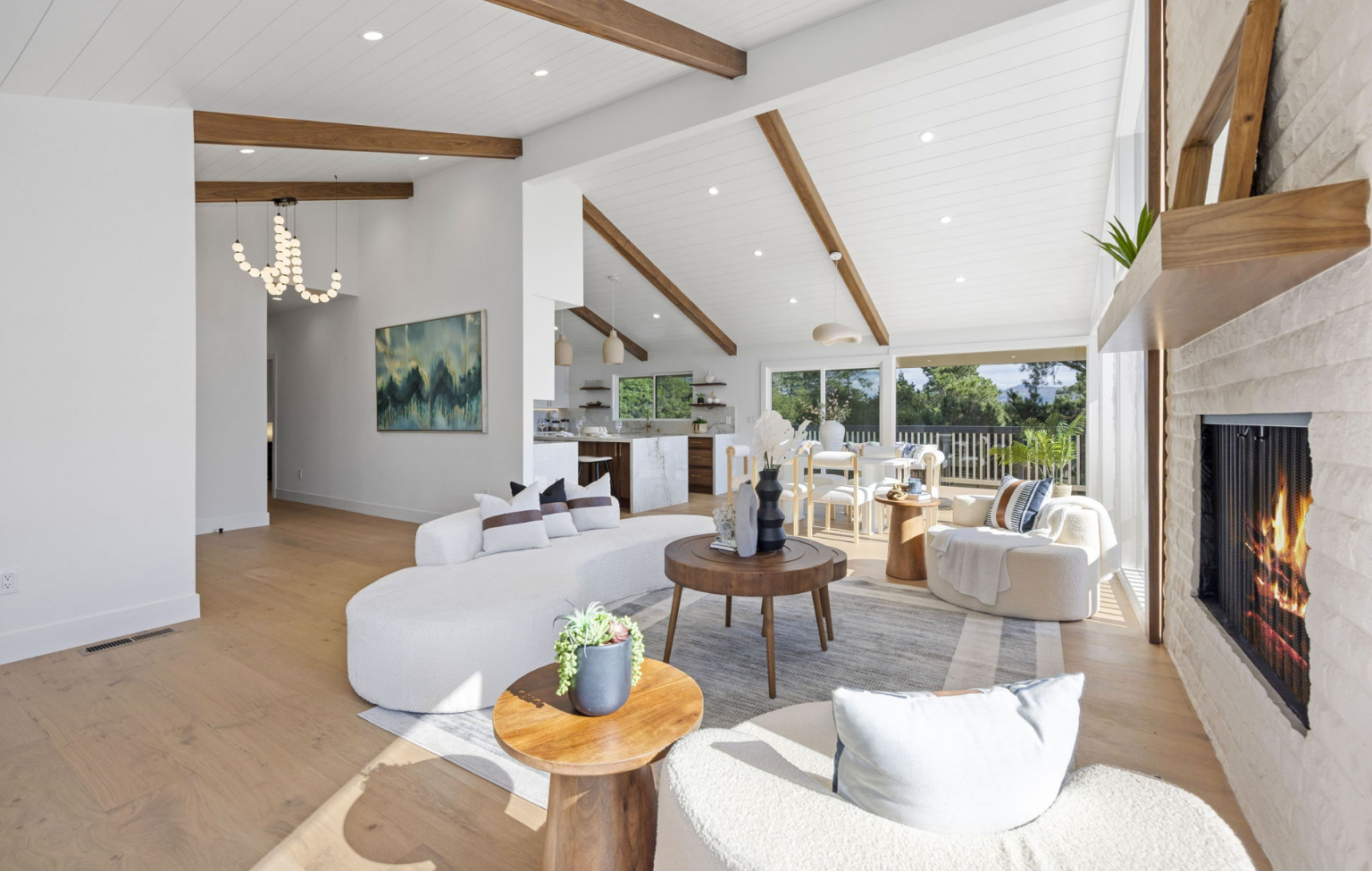 A modern living room with white walls, wood beams, a fireplace, large windows, and curved white sofas highlights this Full Interior-Exterior Home Remodel in Carmel, offering views of the kitchen and lush outdoor greenery.