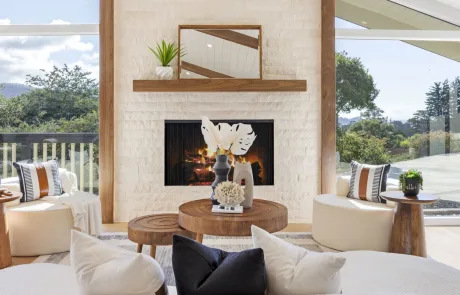 A modern living room from a Full Interior-Exterior Home Remodel in Carmel features a white brick fireplace, wood mantel, circular coffee tables, lush plants, abstract decor, and large windows framing an inviting outdoor view. This photo showcases Bay Area Remodeling Projects.