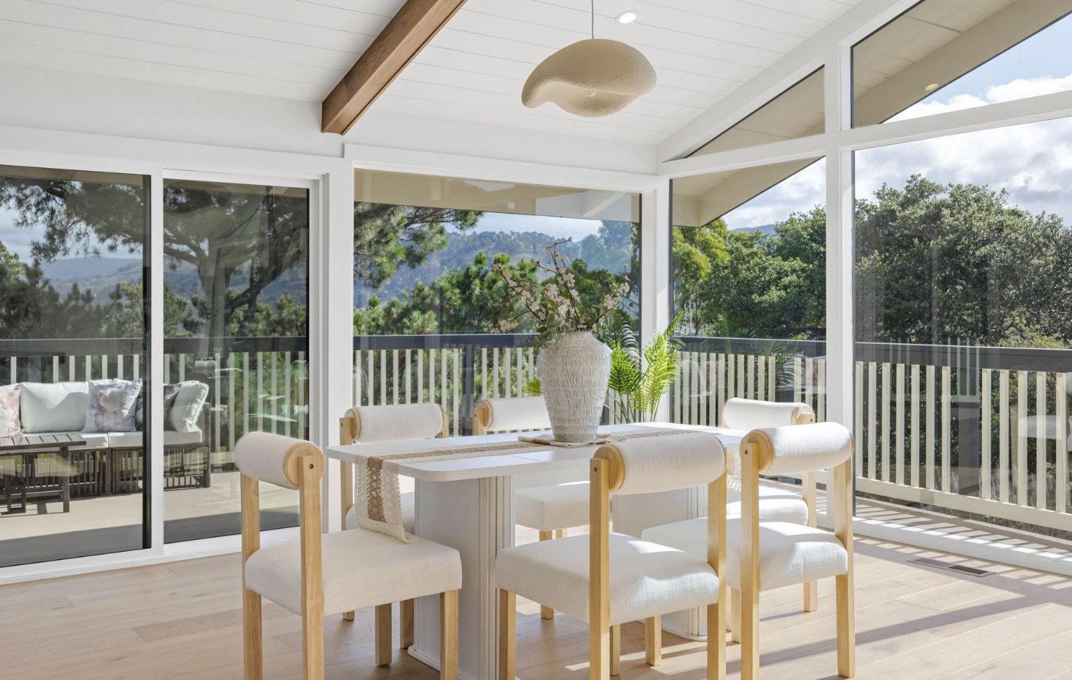 Bright dining area with a white table and six chairs, large windows, glass doors, wooden beams—part of a Full Interior-Exterior Home Remodel in Carmel—opens to an outdoor deck with trees and mountains visible in the background.