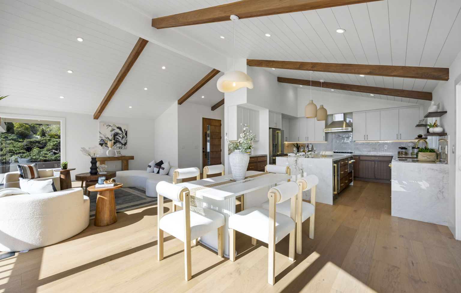 Open-concept living, dining, and kitchen space with wood floors, exposed beams, white walls, and modern furniture under recessed lighting—a highlight of this Full Interior-Exterior Home Remodel in Carmel.
