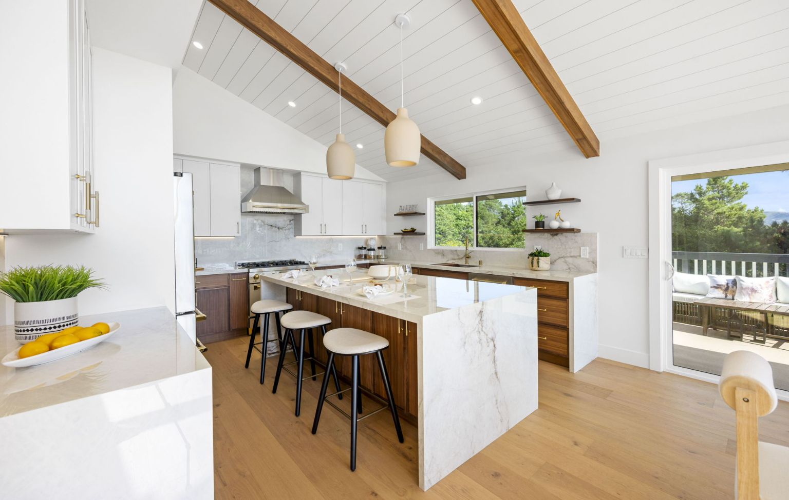 Modern kitchen with white cabinets, marble island with stools, wood accents, pendant lights, and hardwood floors—part of a stunning Full Interior-Exterior Home Remodel in Carmel—features large windows overlooking a deck and trees.