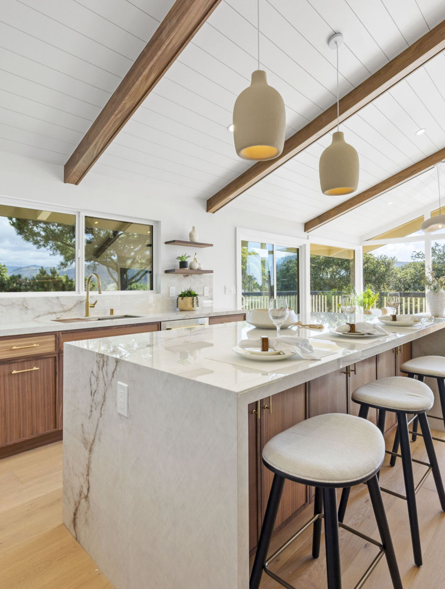 Modern kitchen with marble countertops, wooden cabinets, pendant lights, and bar stools, part of a Full Interior-Exterior Home Remodel in Carmel. Large windows offer stunning views of trees and greenery outside.