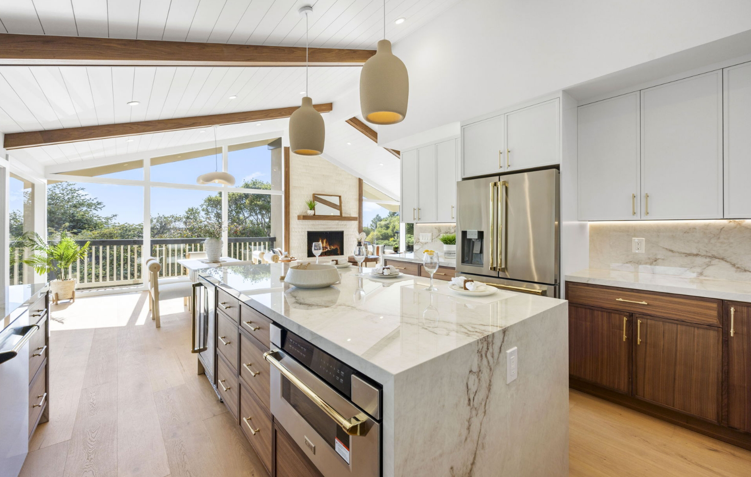 Modern kitchen with a large marble island, pendant lights, wood and white cabinetry, and stainless steel appliances—part of a Full Interior-Exterior Home Remodel in Carmel with an open layout leading to a bright living area with large windows.