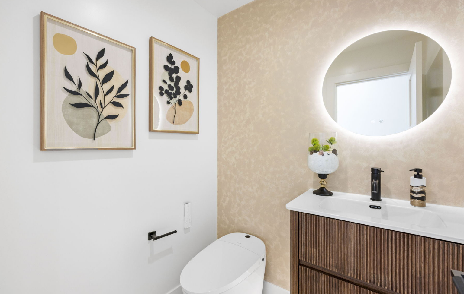 Modern bathroom in a Full Interior-Exterior Home Remodel in Carmel featuring a white toilet, wooden vanity, round backlit mirror, potted plant, and abstract botanical artwork on the wall.