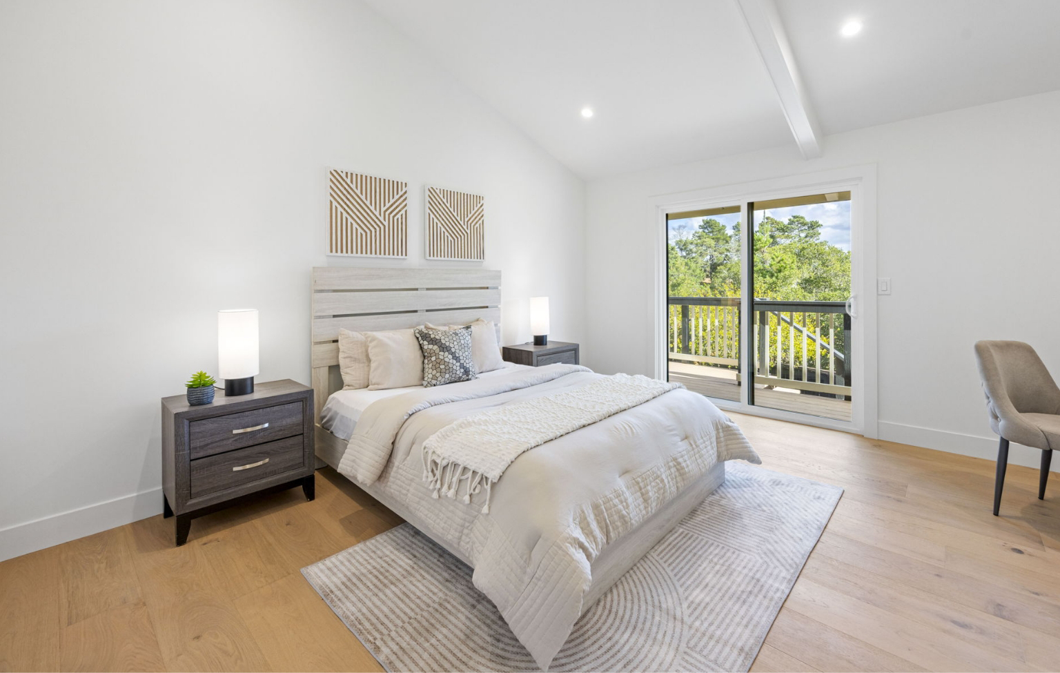 Modern bedroom with a white bed, two nightstands with lamps, a chair, and sliding glass doors opening to a balcony with a view of trees—part of a stunning Full Interior-Exterior Home Remodel in Carmel.