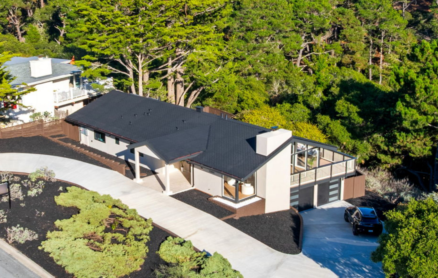 A modern single-story house with a dark roof and large windows, recently updated through a Full Interior-Exterior Home Remodel in Carmel, features a driveway and is surrounded by greenery and trees on a sloped lot.