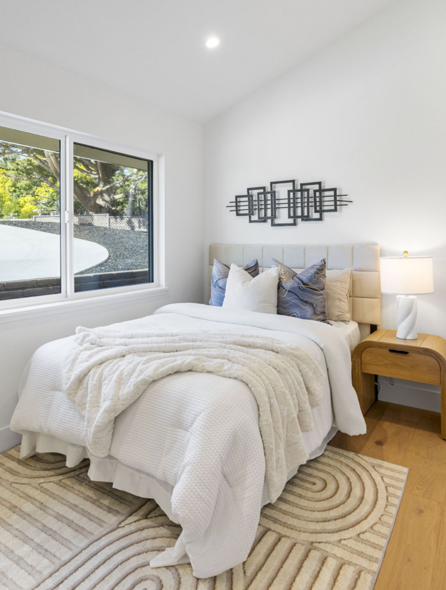 A neatly made bed with white bedding and pillows sits beside a wooden nightstand with a lamp in a bright bedroom, showcasing modern wall art above the headboard—a stunning space from a Full Interior-Exterior Home Remodel in Carmel.