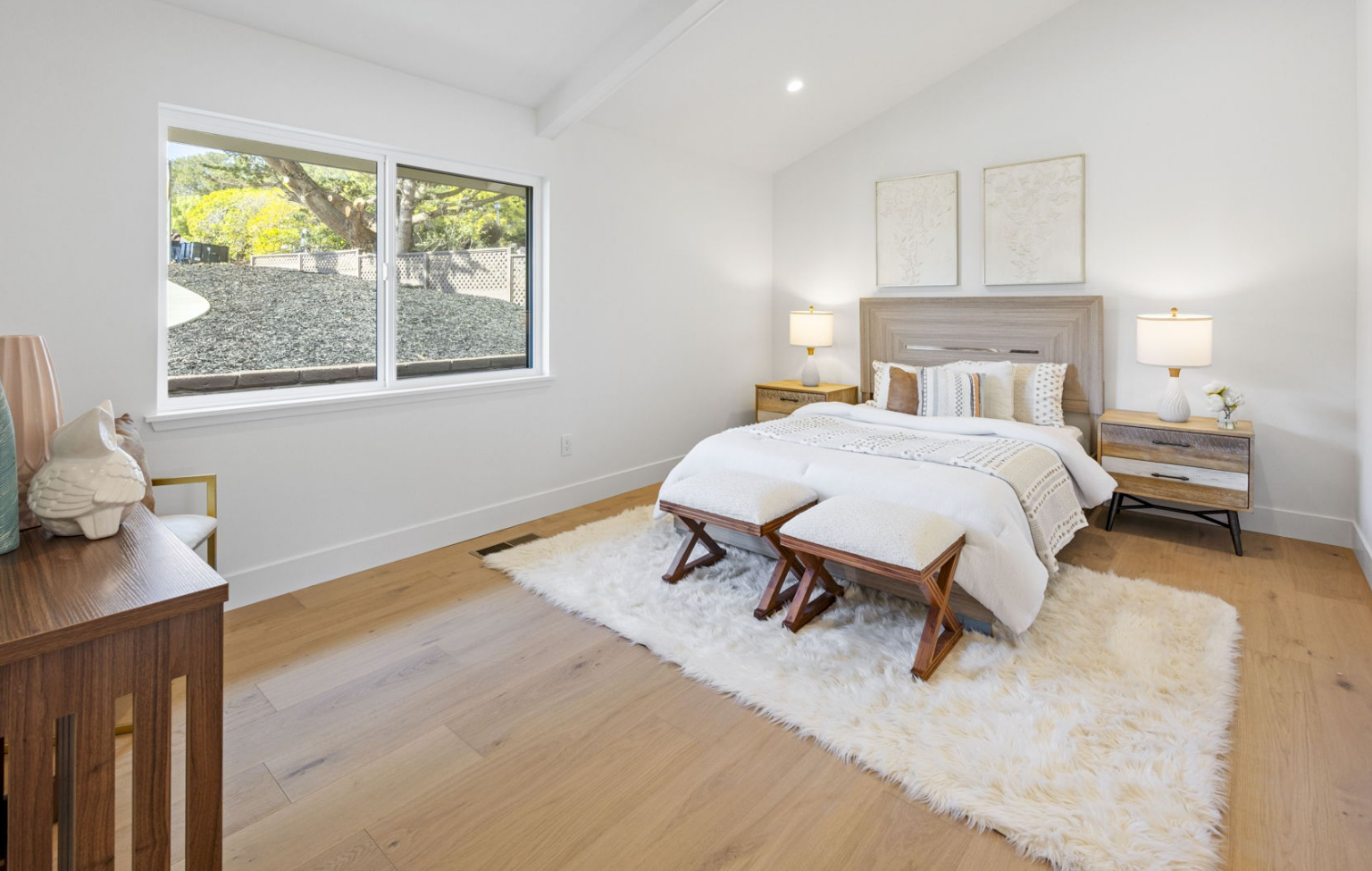 A modern bedroom, part of a Full Interior-Exterior Home Remodel in Carmel, features a double bed, two bedside tables with lamps, a white rug, wooden benches, a window, and minimalistic decor on white walls.