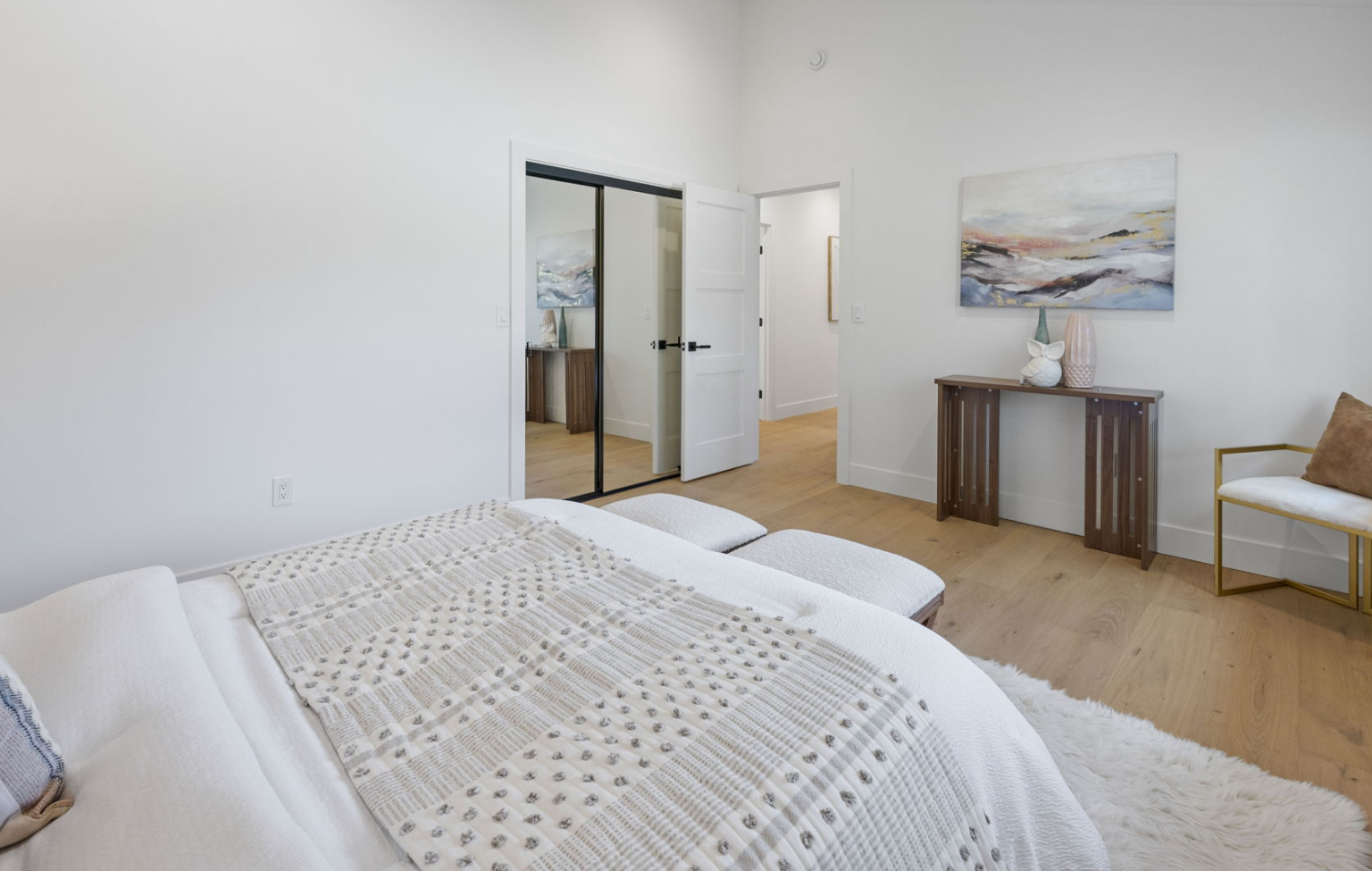 Minimalist bedroom from a Full Interior-Exterior Home Remodel in Carmel, featuring a bed with a patterned blanket, desk with decor, framed art, chair, and light wood flooring. Double glass doors reflect an adjacent room.