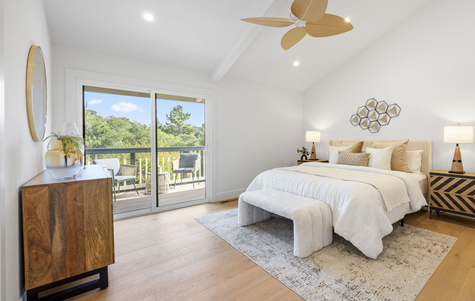 A bright bedroom from a Full Interior-Exterior Home Remodel in Carmel features a large bed, wooden furniture, two lamps, and a sliding glass door leading to a balcony with chairs and views of lush trees.