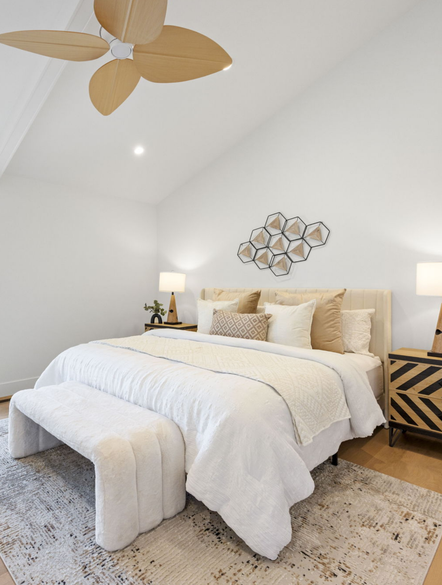 A modern bedroom, part of a Full Interior-Exterior Home Remodel in Carmel, features a large bed with neutral bedding, two bedside tables with lamps, geometric wall art, and a ceiling fan with wooden blades.