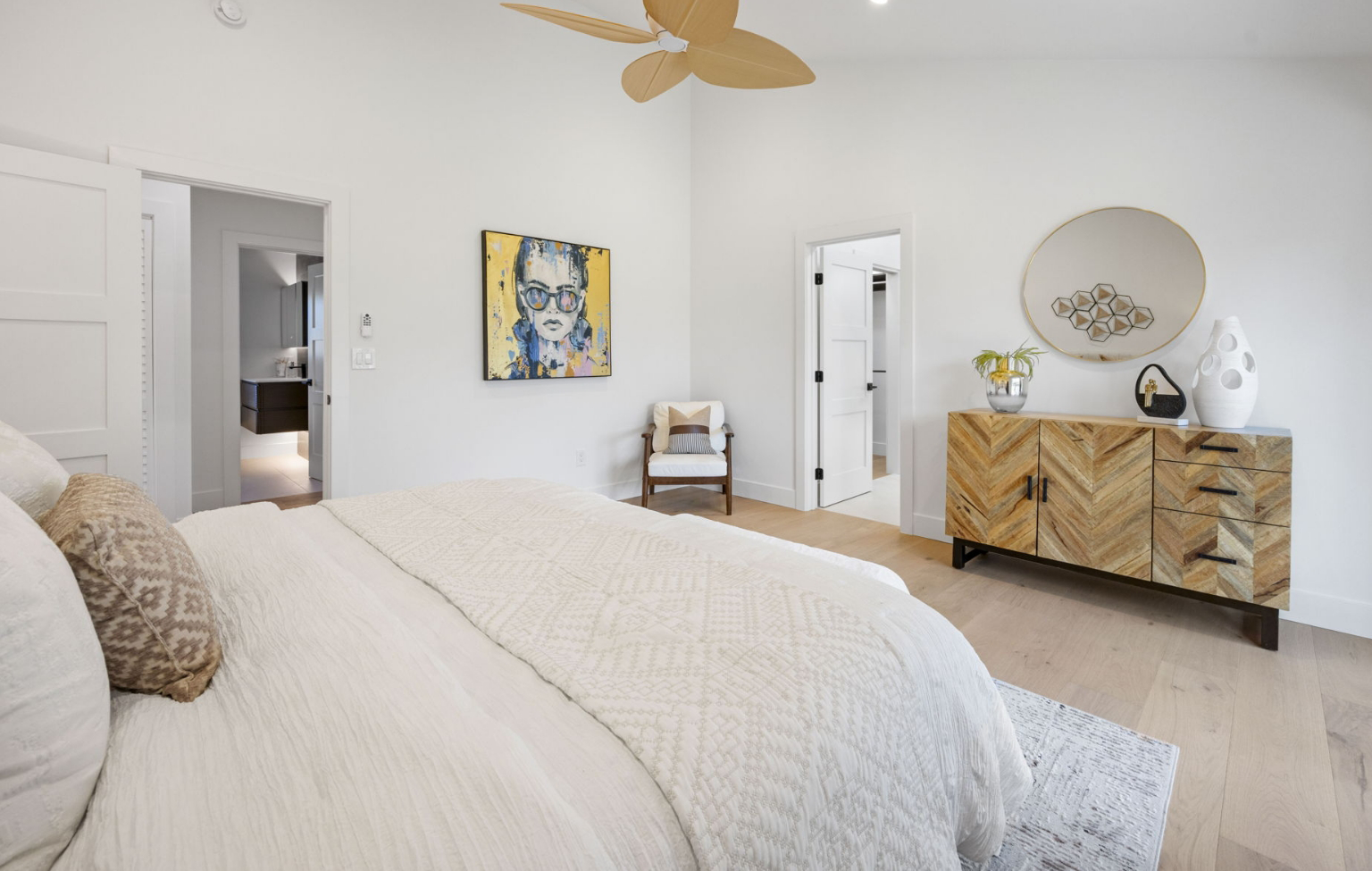 Modern bedroom from a Full Interior-Exterior Home Remodel in Carmel, featuring light wood floors, a large bed with white bedding, a dresser with a round mirror, abstract wall art, and a ceiling fan with leaf-shaped blades.