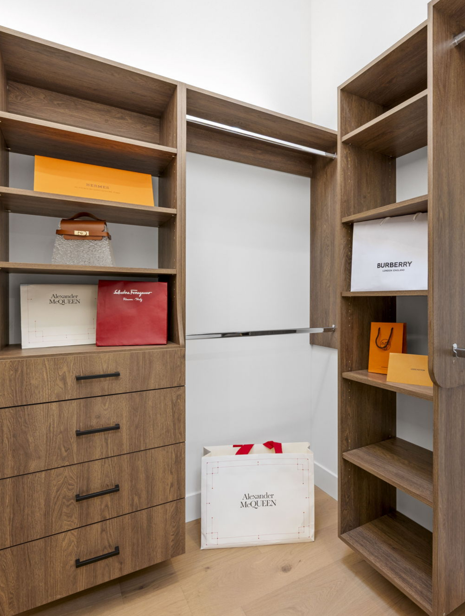 A modern walk-in closet with wood shelves and drawers, part of a Full Interior-Exterior Home Remodel in Carmel, features shopping bags from luxury brands like Alexander McQueen, Burberry, Hermès, and Salvatore Ferragamo.