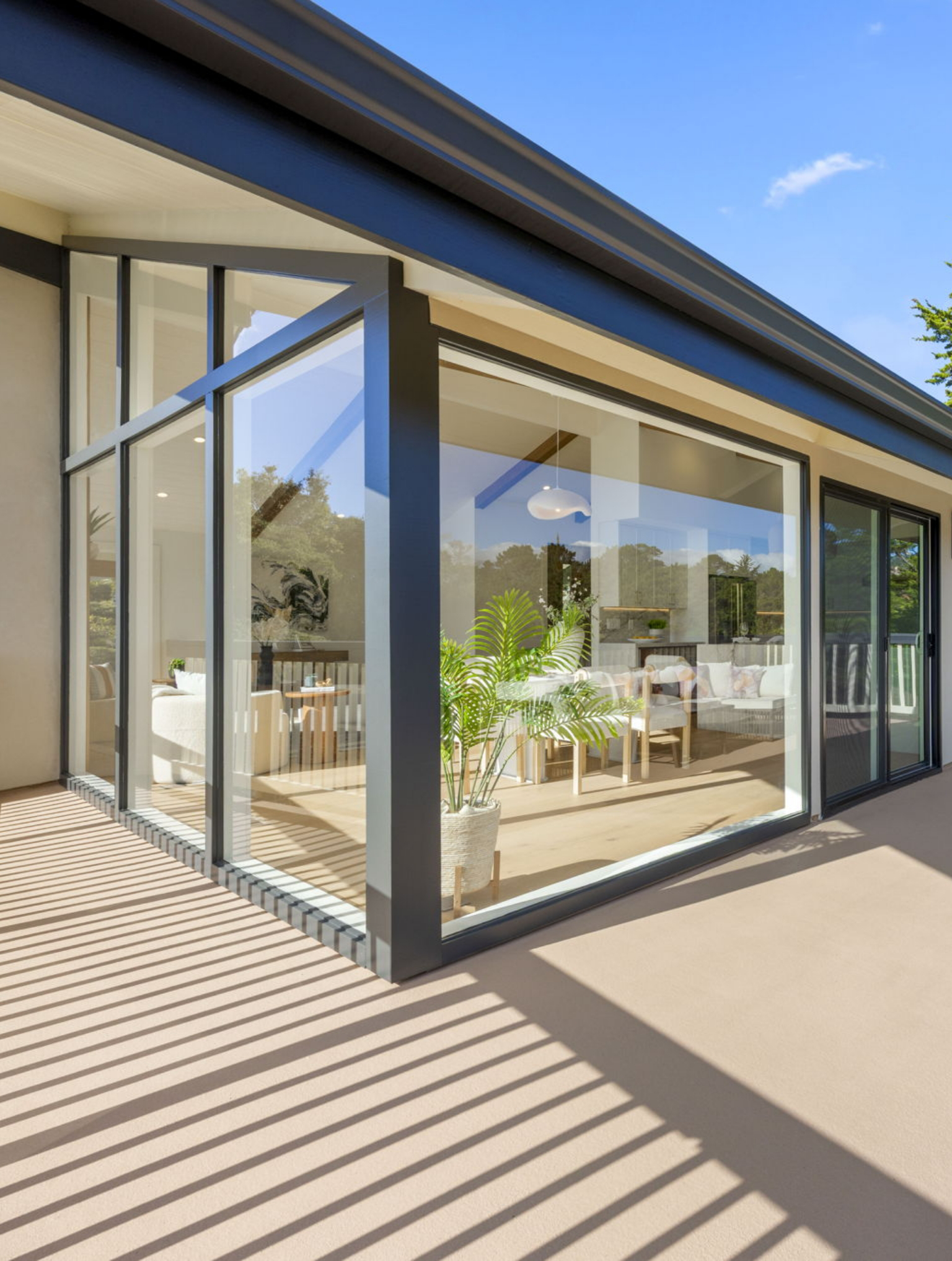 A modern house with large floor-to-ceiling windows, a visible dining area, and a potted plant near the glass showcases this Full Interior-Exterior Home Remodel in Carmel under clear blue skies.