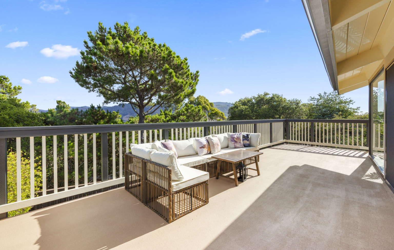A spacious outdoor patio with cushioned seating and a coffee table, part of a Full Interior-Exterior Home Remodel in Carmel, is surrounded by a wooden railing and overlooks trees and distant hills under a clear sky.