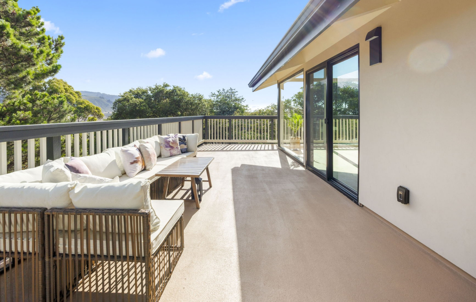 Spacious outdoor patio with cushioned seating, coffee tables, and railing, adjacent to a modern building—part of a Full Interior-Exterior Home Remodel in Carmel—with large glass doors and scenic views of trees and hills.