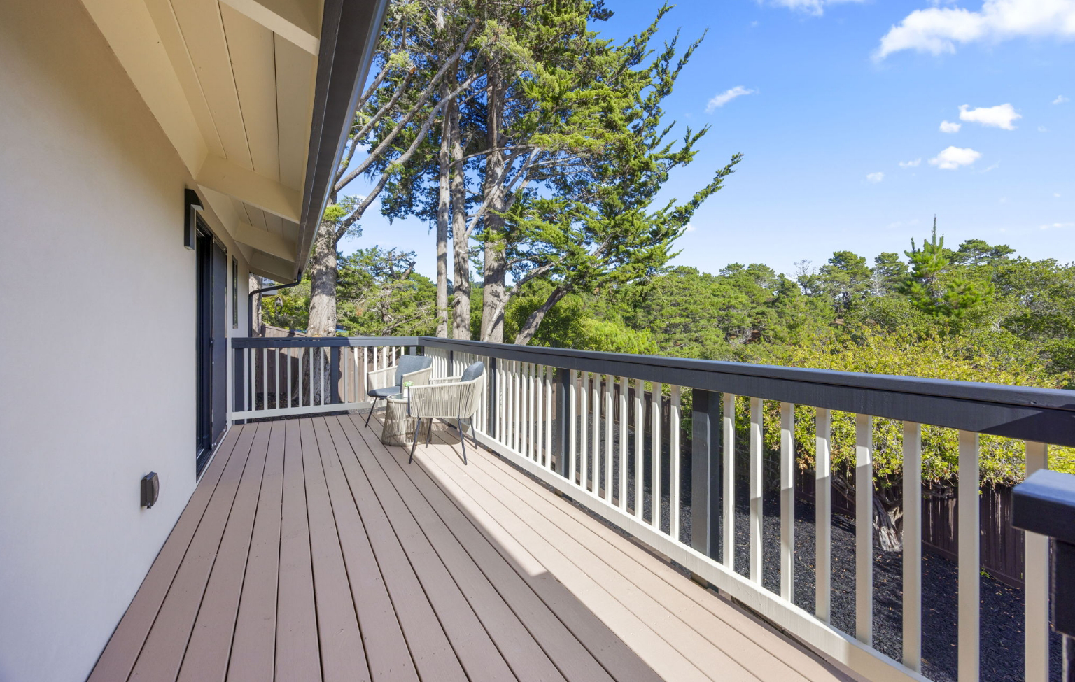 A wooden balcony with black and white railing, two chairs, and a small table overlooks trees under a clear blue sky—part of a Full Interior-Exterior Home Remodel in Carmel.