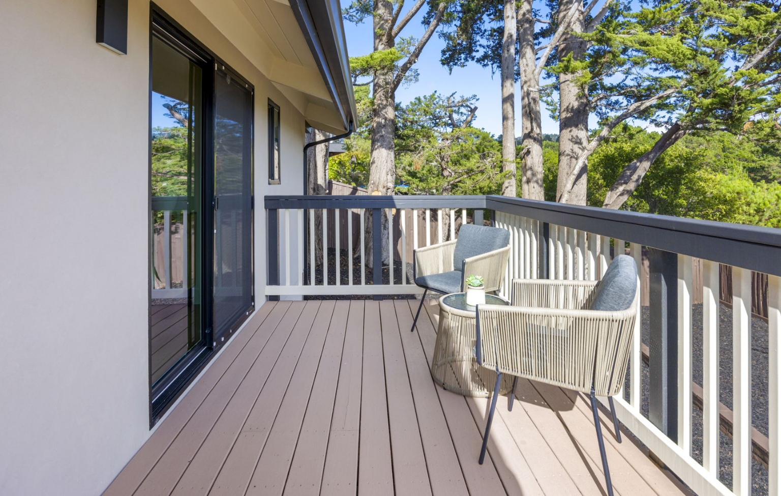 A small balcony from a Full Interior-Exterior Home Remodel in Carmel features two chairs, a round side table, and a railing that overlooks trees on a sunny day.