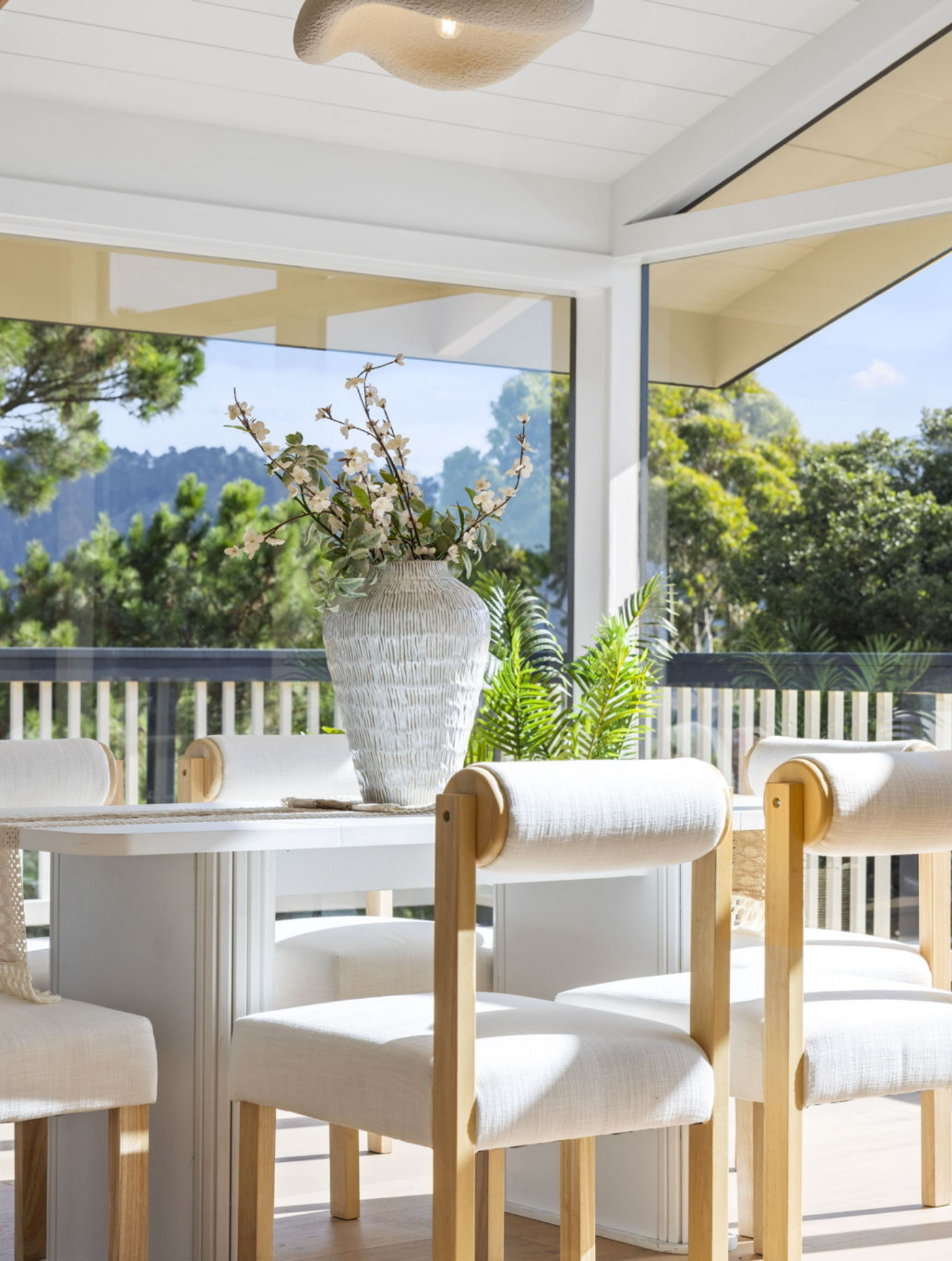 A round white table with a woven vase of flowers is surrounded by light wood and upholstered chairs on a bright, covered patio—part of a Full Interior-Exterior Home Remodel in Carmel, with lush greenery visible just outside.