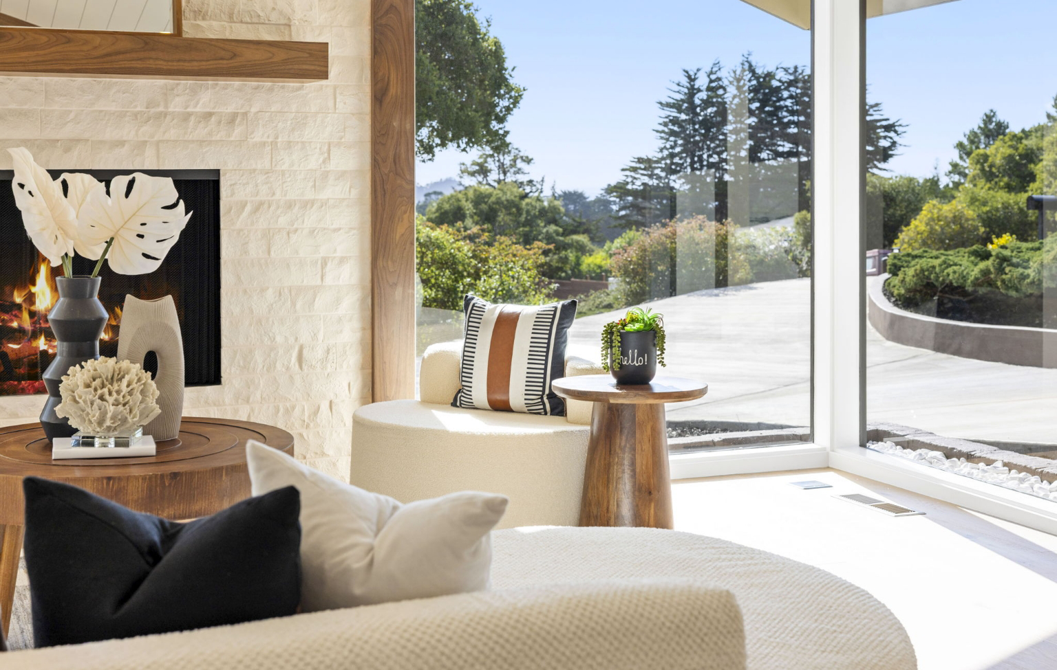 Modern living room with large windows, light-colored furniture, a fireplace, decorative pillows, and a small side table with a potted plant highlight this Full Interior-Exterior Home Remodel in Carmel. Trees and driveway are visible outside.