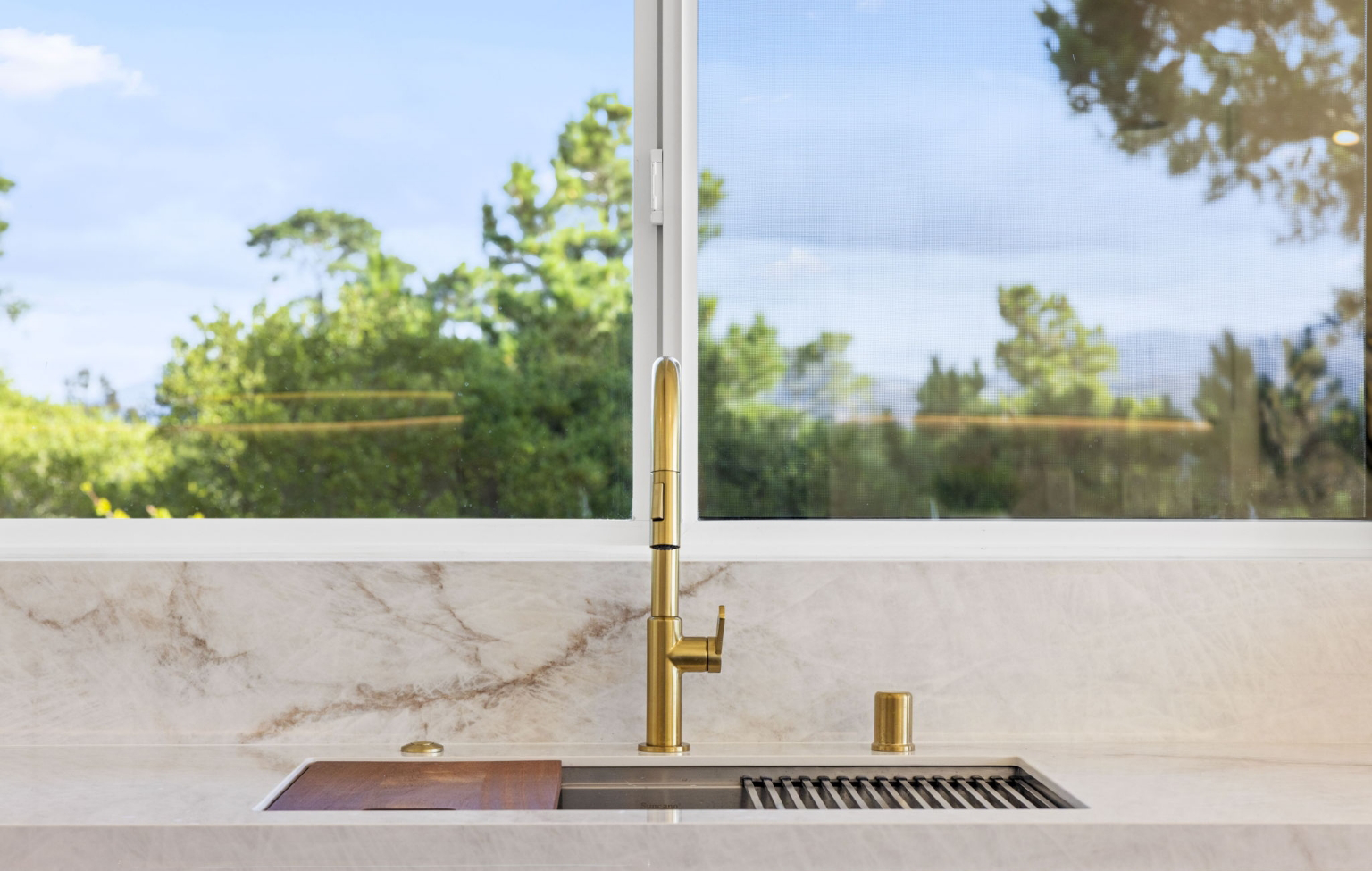 A modern kitchen sink with a gold faucet is set in a marble countertop, positioned in front of a large window overlooking greenery—a highlight of this Full Interior-Exterior Home Remodel in Carmel.