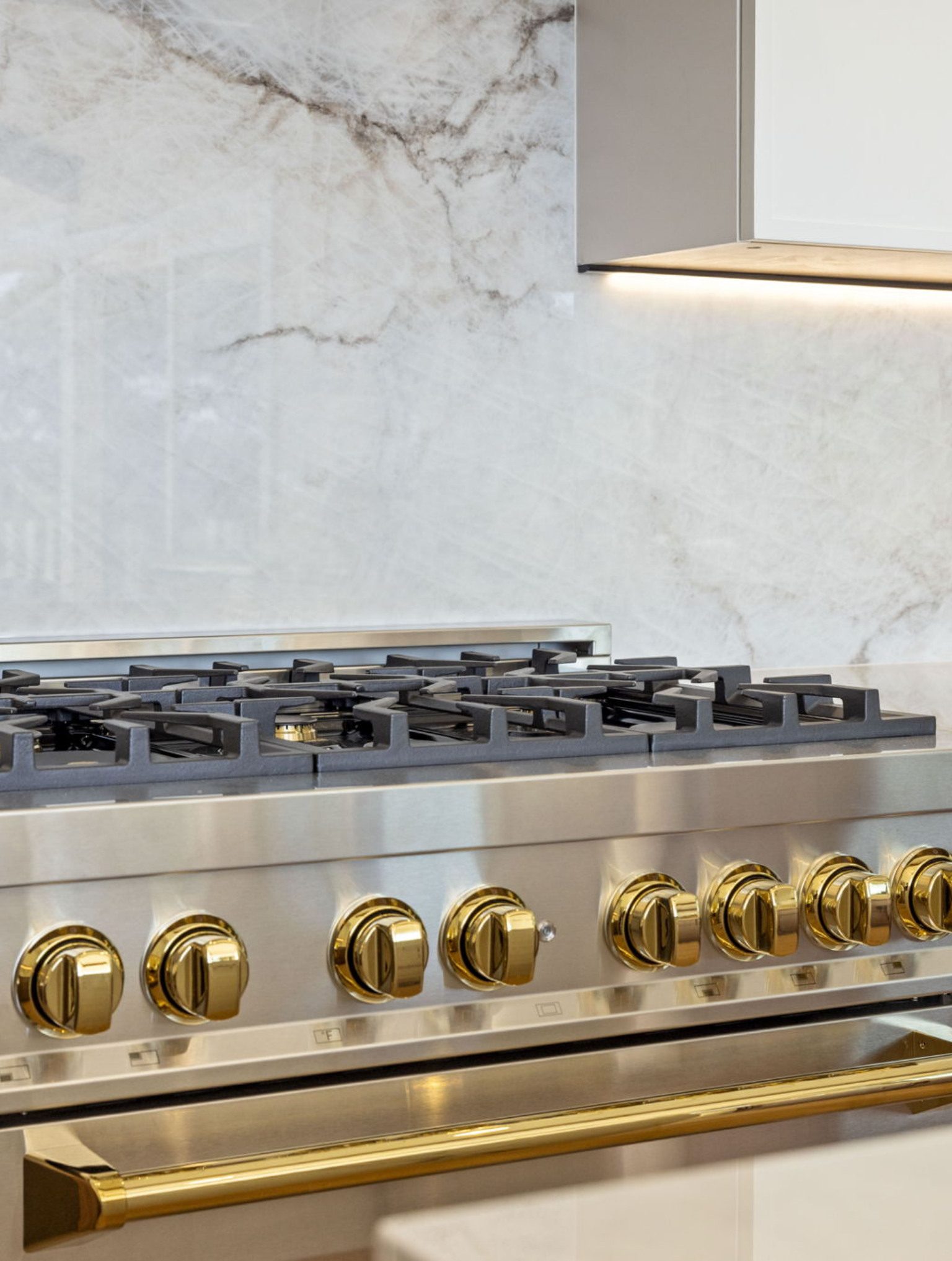 A close-up of a stainless steel gas stove with gold-colored knobs and accents, set against a marble backsplash in a modern kitchen from a Full Interior-Exterior Home Remodel in Carmel.