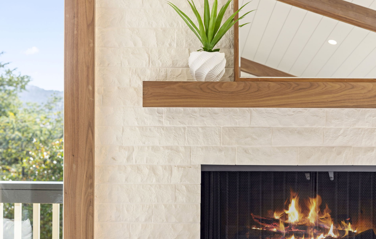 Close-up of a modern fireplace with a burning fire, cream stone surround, and wooden mantel—part of a Full Interior-Exterior Home Remodel in Carmel—with a potted plant in a white ceramic vase on top.