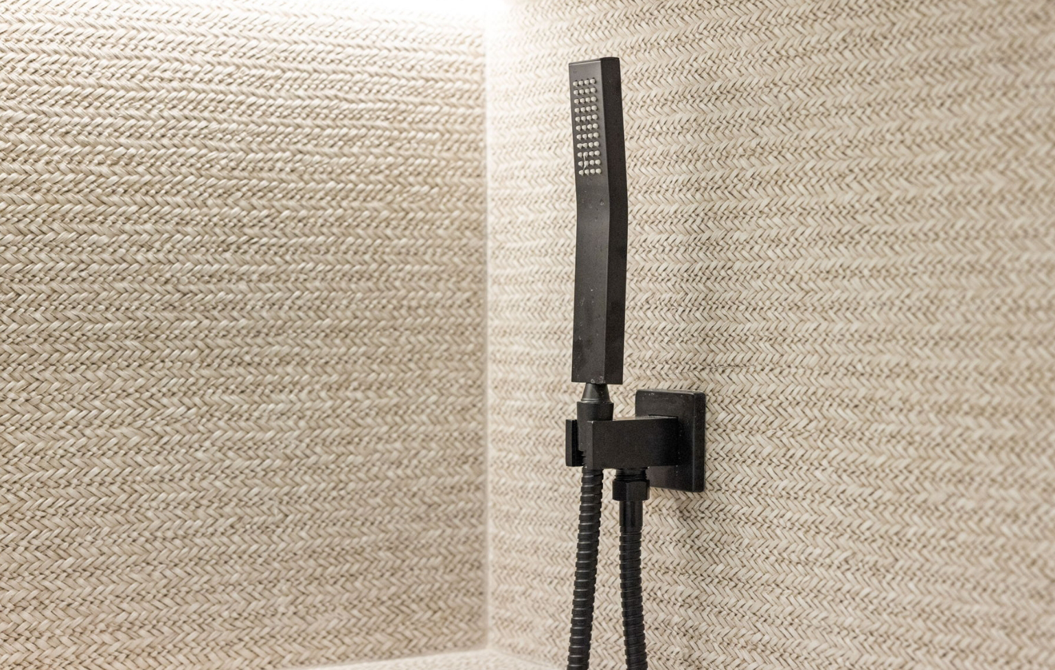 A modern black handheld showerhead is mounted on a textured beige herringbone-patterned wall in a bathroom corner, part of a Full Interior-Exterior Home Remodel in Carmel.