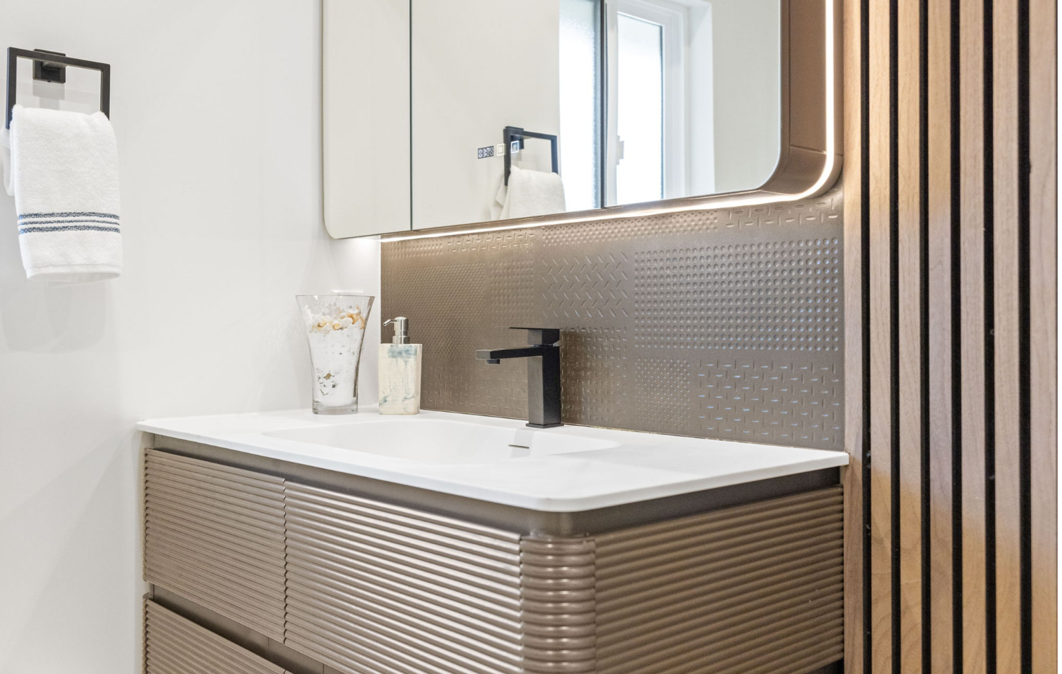 Modern bathroom vanity with a white countertop, black faucet, soap dispenser, glass jar, wall mirror, towel ring, and vertical wooden slats on the right wall—part of a Full Interior-Exterior Home Remodel in Carmel.