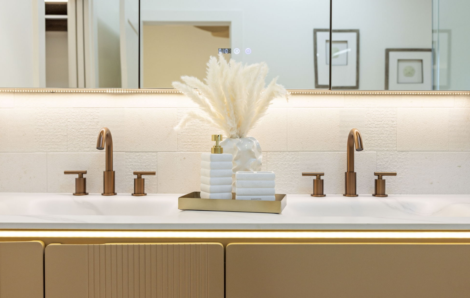 A modern bathroom countertop from a Full Interior-Exterior Home Remodel in Carmel features two bronze faucets, a decorative vase with pampas grass, stacked towels, and a soap dispenser on a gold tray, all set against a large mirror.