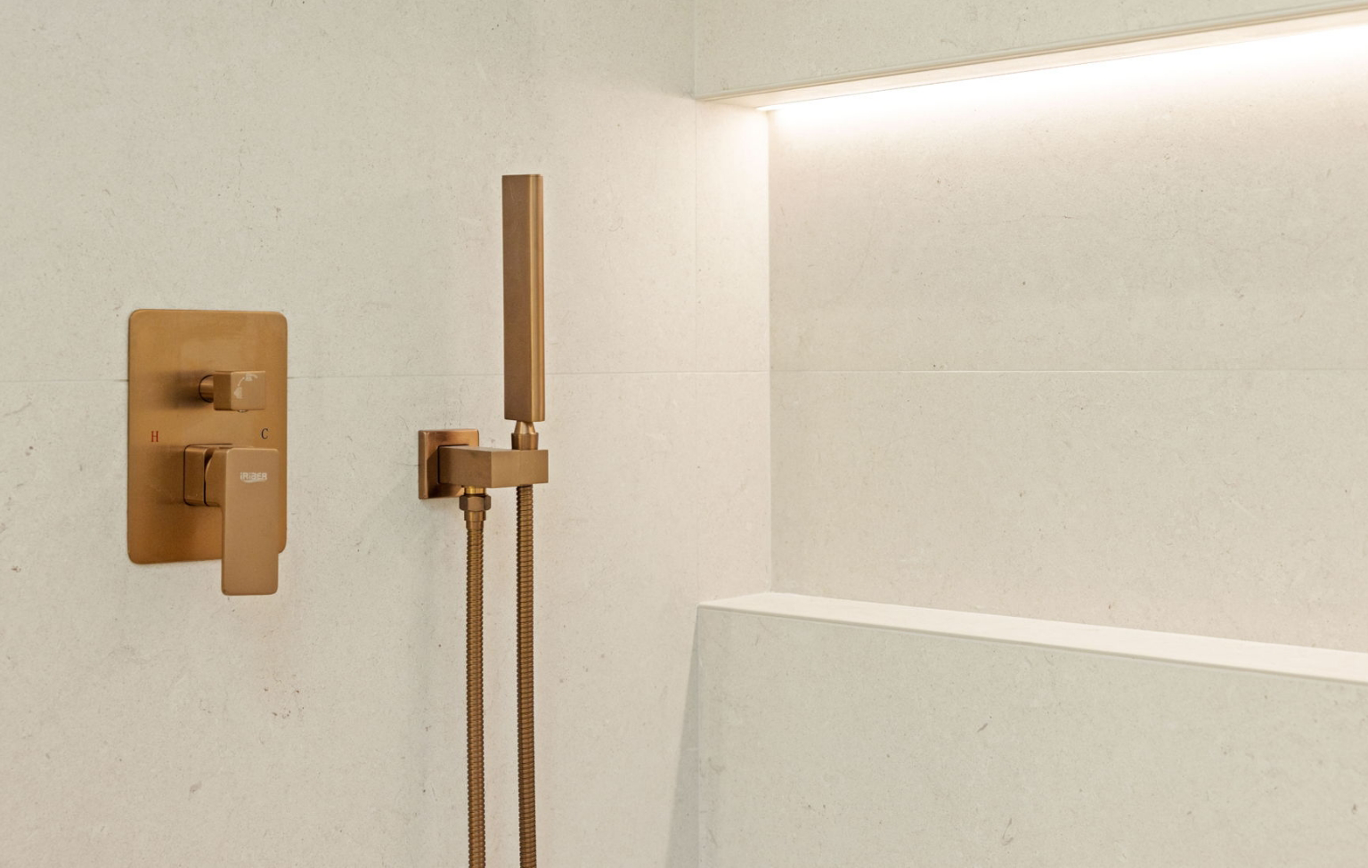 A modern gold shower fixture with handheld sprayer is mounted on a light beige tiled wall with a recessed shelf and LED lighting above—part of a stunning Full Interior-Exterior Home Remodel in Carmel.