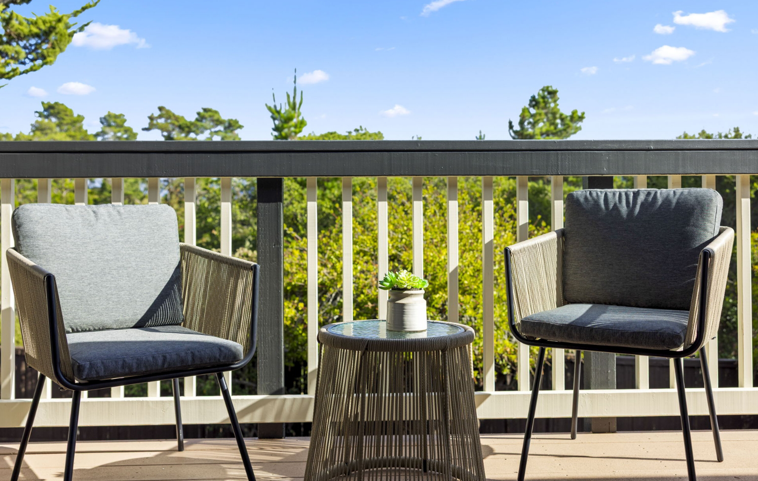 Two cushioned patio chairs and a small round table with a potted succulent are set on a balcony overlooking trees and a clear blue sky—a perfect spot to relax after a Full Interior-Exterior Home Remodel in Carmel.