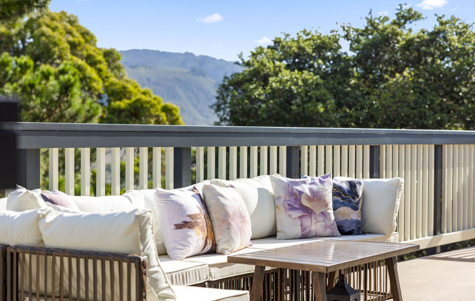 Outdoor patio with cushioned seating and decorative pillows beside a wooden table, set against a railing overlooking green trees and distant hills—part of a Full Interior-Exterior Home Remodel in Carmel under a blue sky.