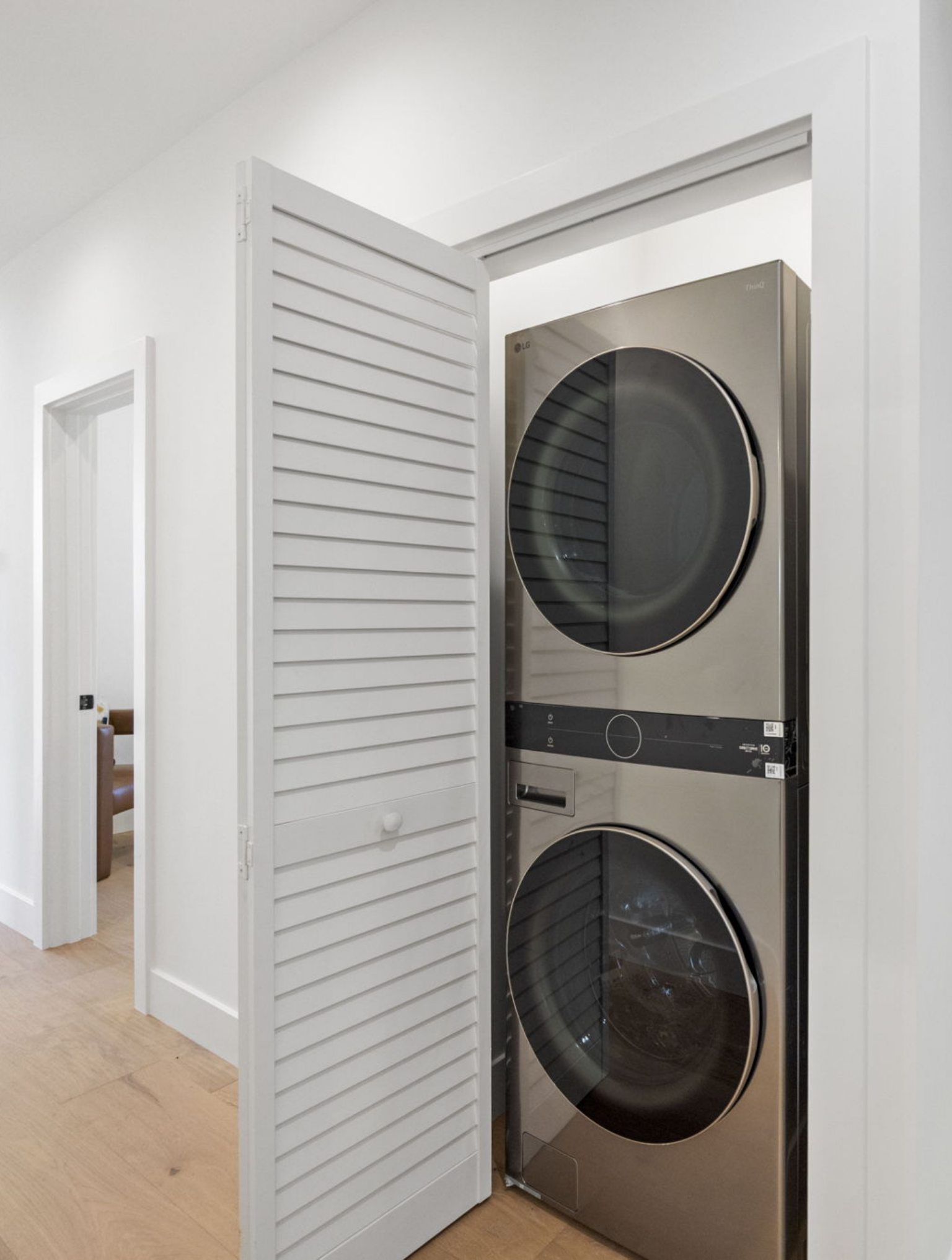 A stacked washer and dryer set is installed in a small laundry closet with a white louvered door, part of a Full Interior-Exterior Home Remodel in Carmel, in a hallway with light wood flooring.