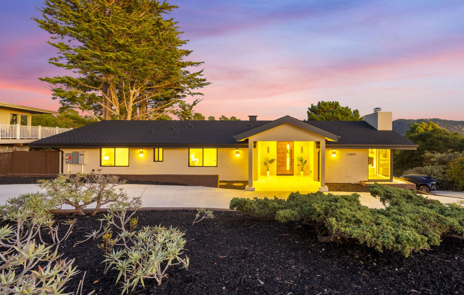 Single-story modern house featuring a Full Interior-Exterior Home Remodel in Carmel, with large windows and a well-lit entrance at dusk, surrounded by a landscaped yard with plants and trees.