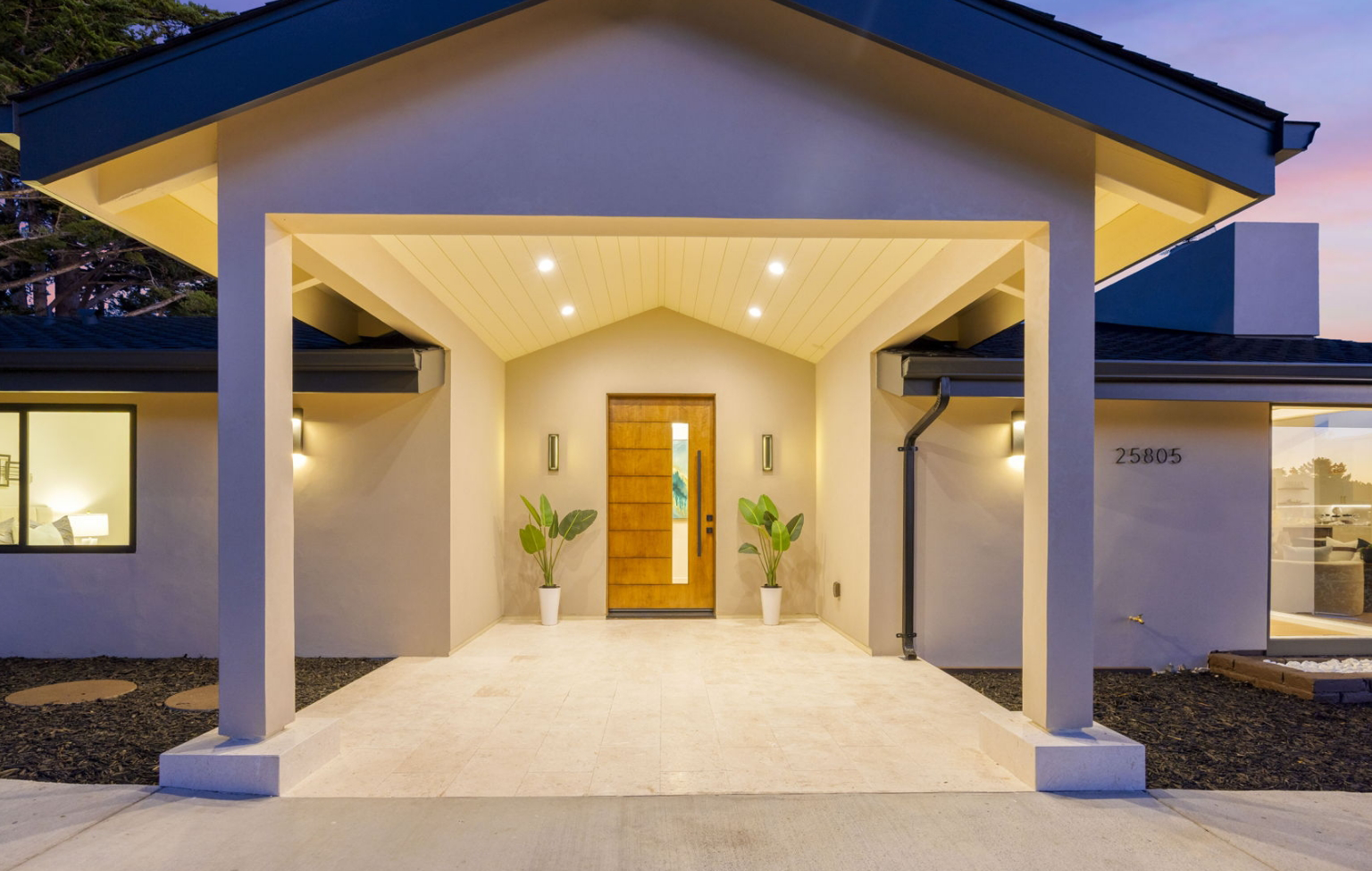 Modern house entrance with a wood door, beige walls, two potted plants, and recessed ceiling lights, photographed at dusk. This inviting entryway is part of a Full Interior-Exterior Home Remodel in Carmel. House number 25805 is visible on the right wall.