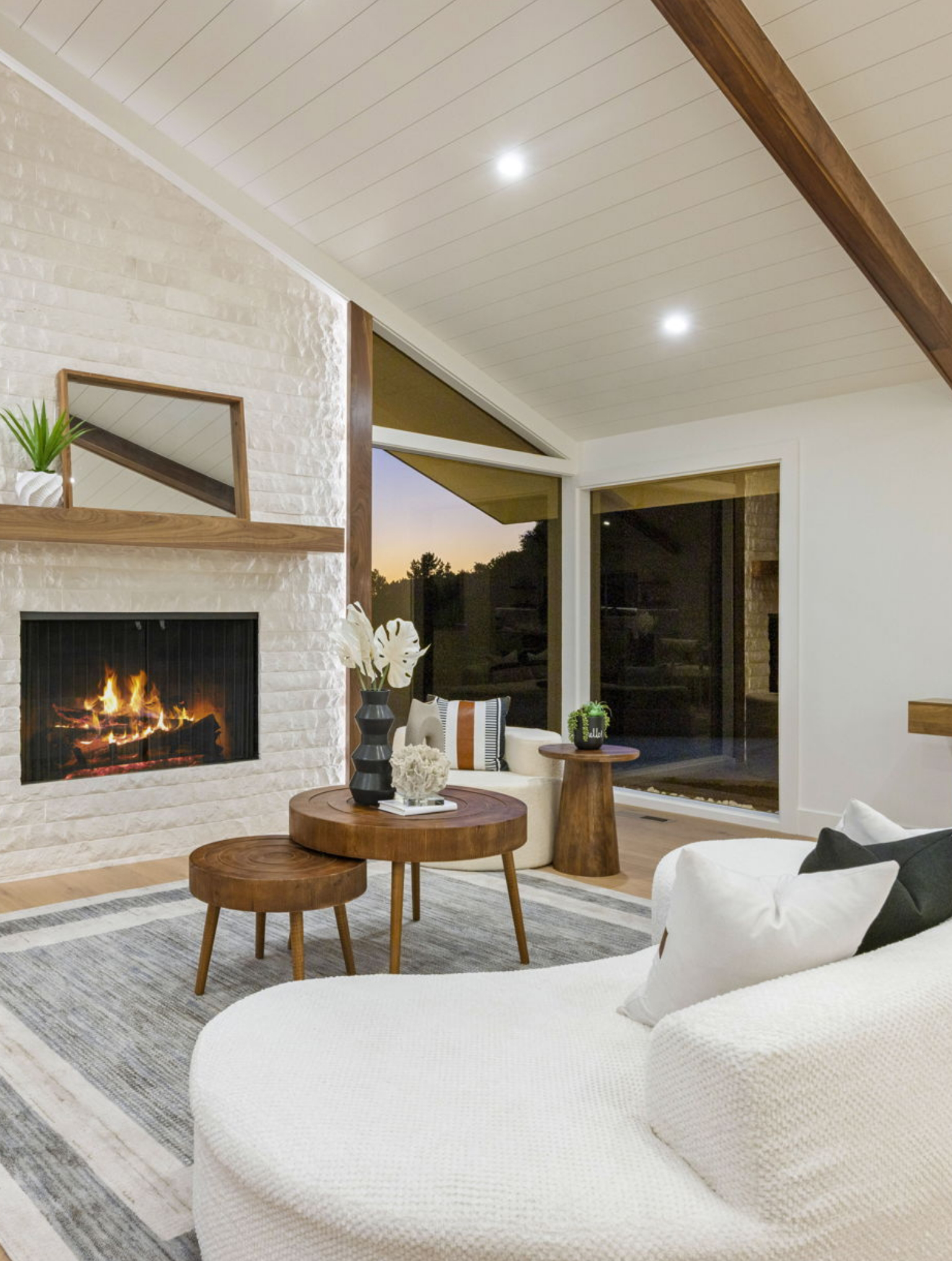 Modern living room with a white brick fireplace, burning fire, wooden ceiling beams, round wooden tables, a white curved sofa, and large windows showcasing a Full Interior-Exterior Home Remodel in Carmel at sunset.