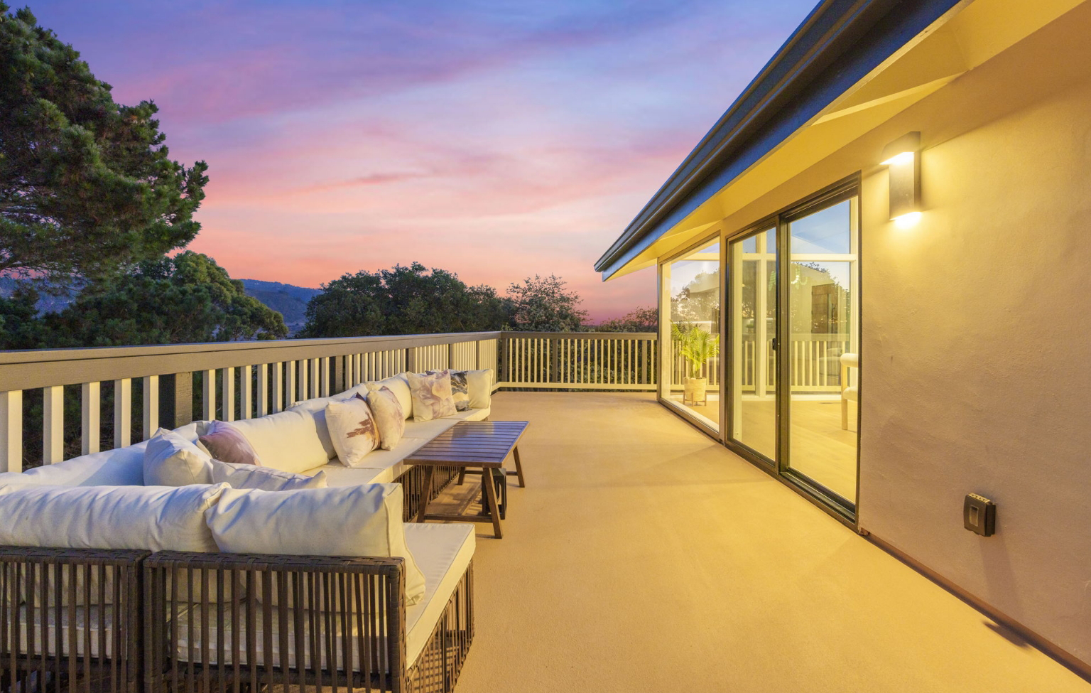 Spacious outdoor patio with cushioned seating, coffee tables, and railing, adjoining a modern house at sunset with trees in the background—a stunning result of a Full Interior-Exterior Home Remodel in Carmel.