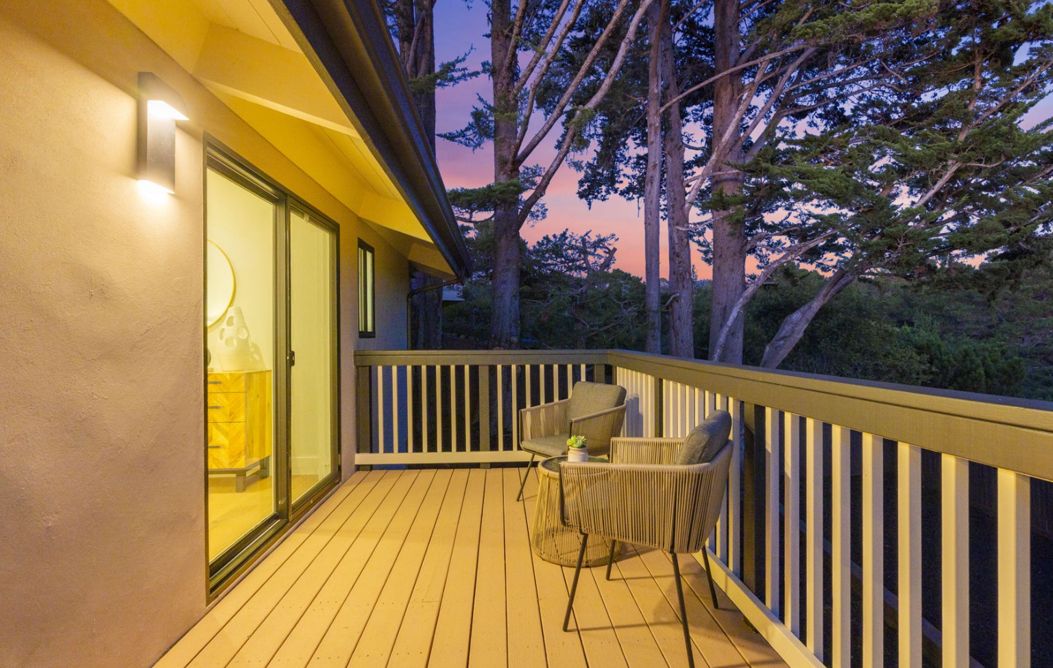 A wooden balcony with two chairs and a small table overlooks trees at sunset. Perfect after a Full Interior-Exterior Home Remodel in Carmel, the space features a glass door leading inside and a wall light mounted beside it.
